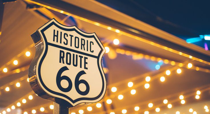 A white sign reads "Historic Route 66" in black ink. String lights draping from the ceiling of a structure are in the background.