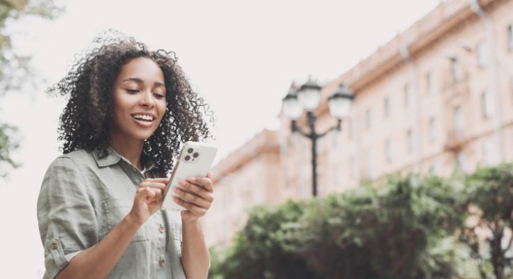 A smiling woman is standing outside near a large building, looking at her iPhone. Trees are in the background.