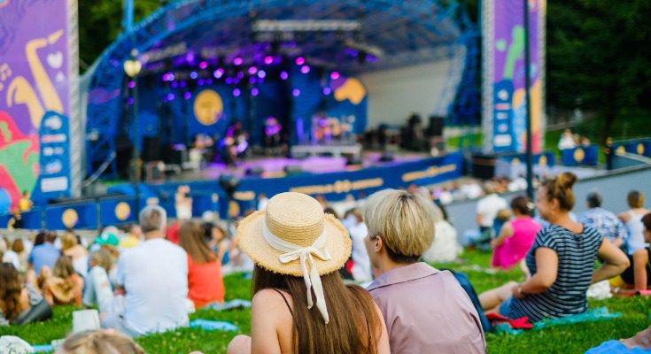Two people sit together on the grass and watch a colorful concert stage as a crowd relaxes around them.