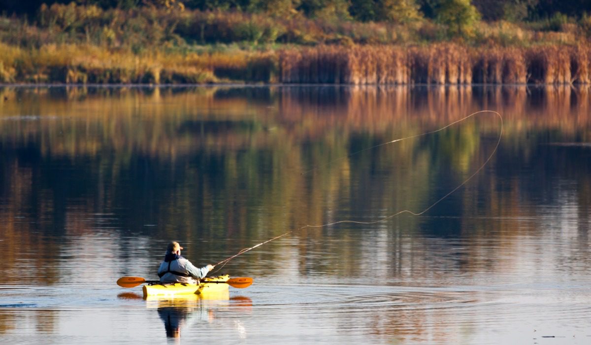 A person is fishing from a yellow kayak on a calm lake. The trees in the background are turning orange.