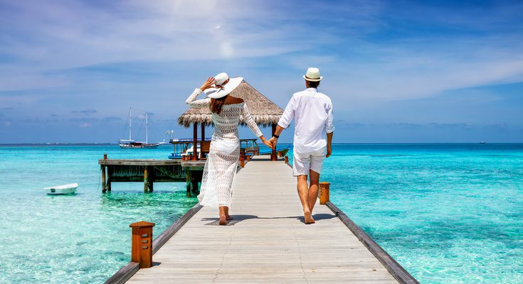 A newlywed couples takes a walk holding hands down a boardwalk that hovers over clear blue waters. A hut waits at the end.