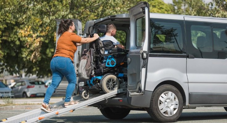 A woman in an orange shirt and jeans pushes a man in an electronic wheelchair up a ramp into the back of a van.