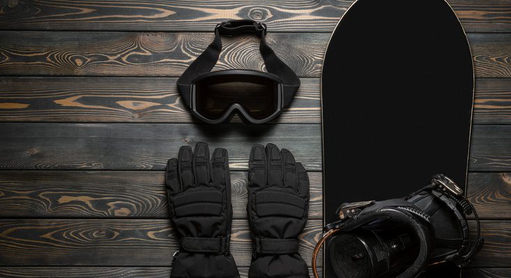 A black snowboard, black snowboarding goggles, black gloves, and black boots sit on a wooden surface.