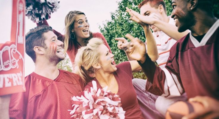 Football fans wearing team colors cheer and celebrate together at an outdoor tailgate.