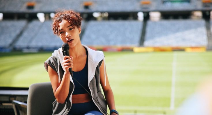 A person wearing a sleeveless hoodie holds a microphone in front of a football field on a bright, sunny day.