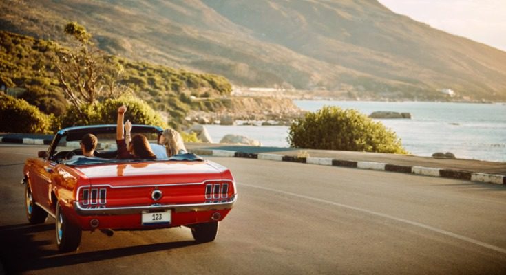 Three people riding in a red convertible along a coastal road with mountains and the ocean in the background.