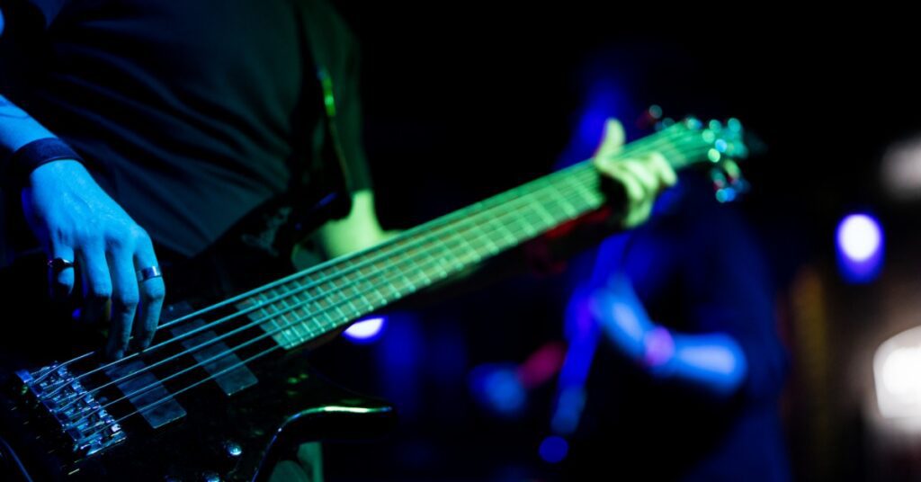 A close-up of a black electric guitar that a man in a black shirt is playing on a stage with blue and green spotlights.
