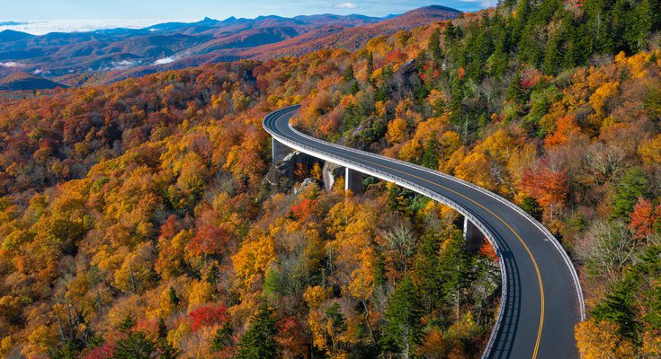 A winding two-lane road winds around a mountain that is surrounded by trees that display red, orange, and yellow leaves.
